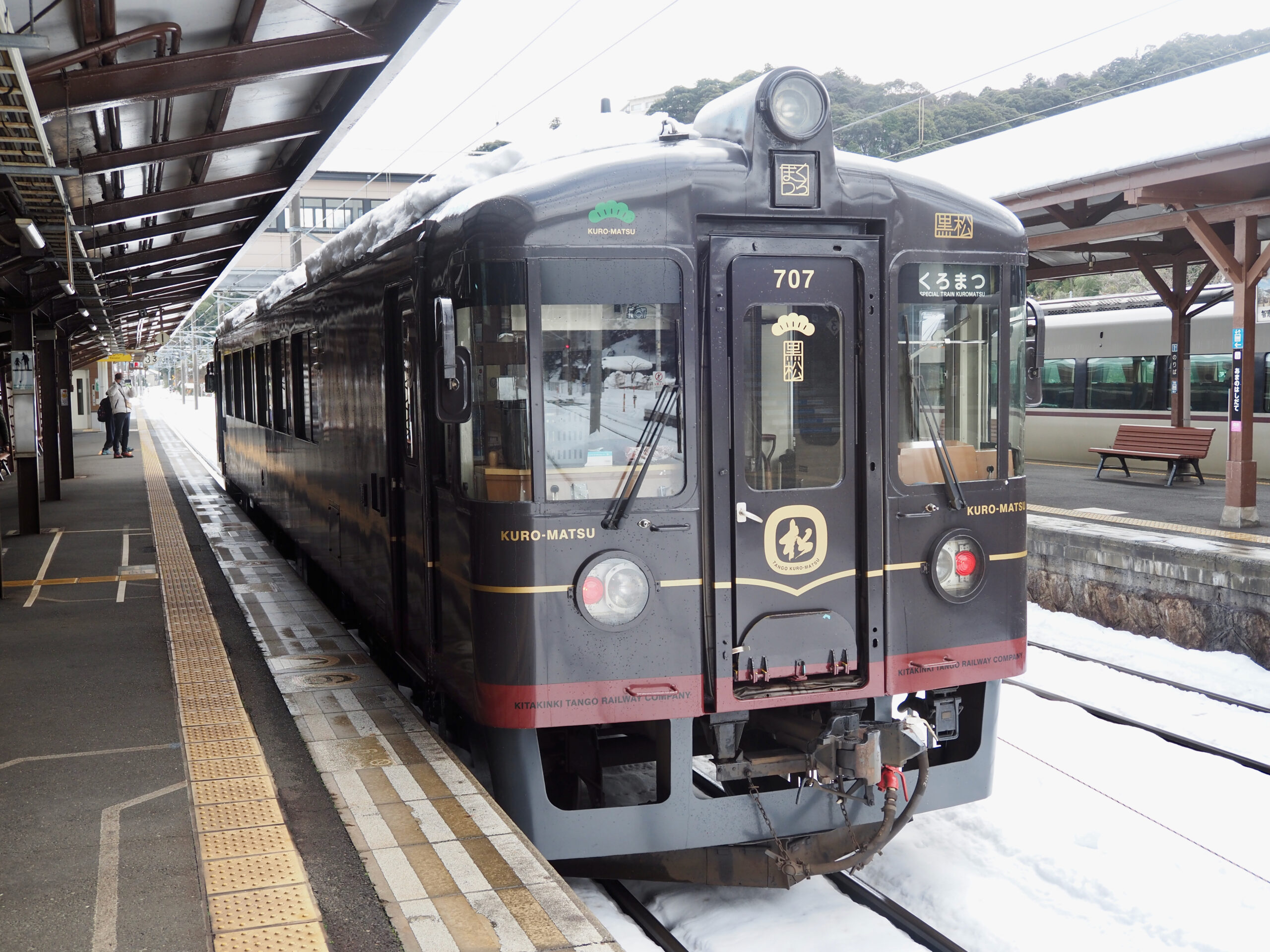 black Kuro-Matsu train is stationed at a snowy platform, with snow visible on the train roof and tracks. A few people are seen on the platform, bundled up against the cold.