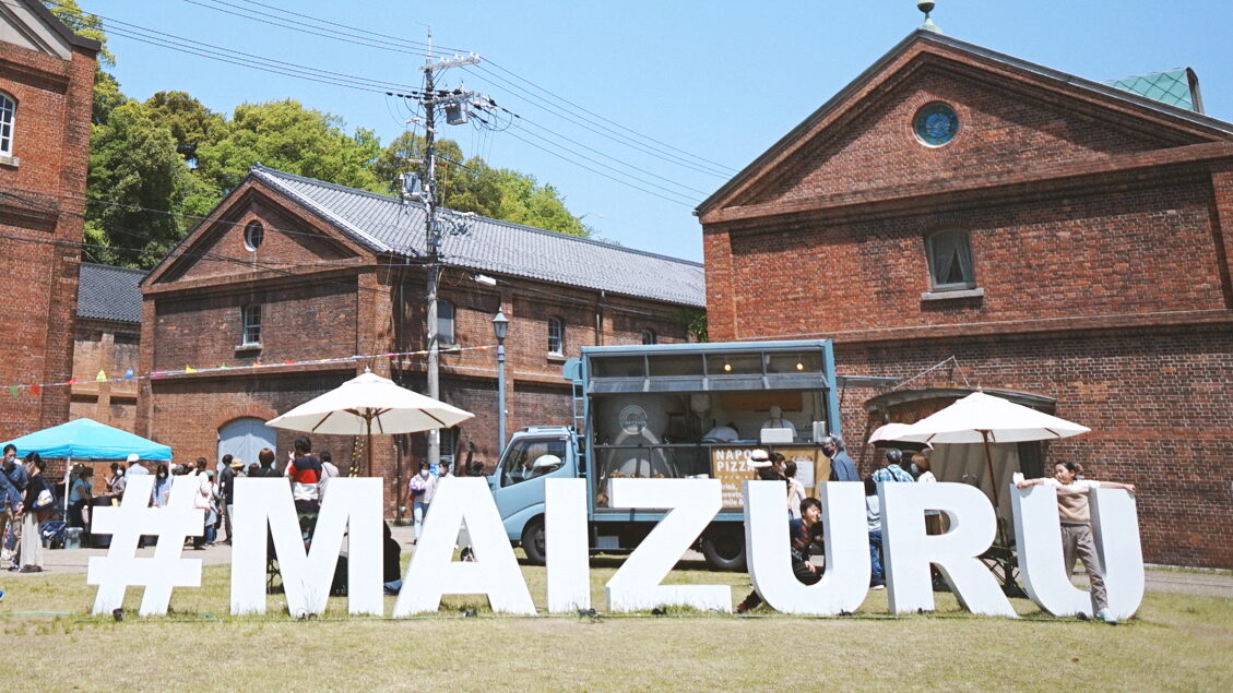 A close-up shot of the #Maizuru sign at the Maizuru Red Brick Park with food trucks and people at tables behind it